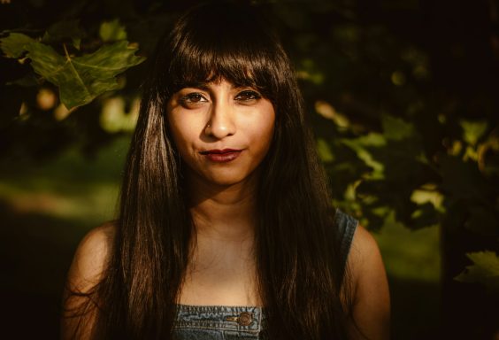 A young woman with long dark hair stands outdoors among green foliage.