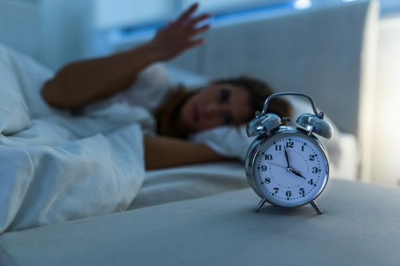 A person in bed looks alarmed as a silver alarm clock sits nearby.