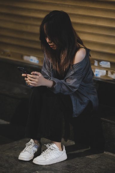 A young woman sits on the ground, looking at her phone, dressed casually.