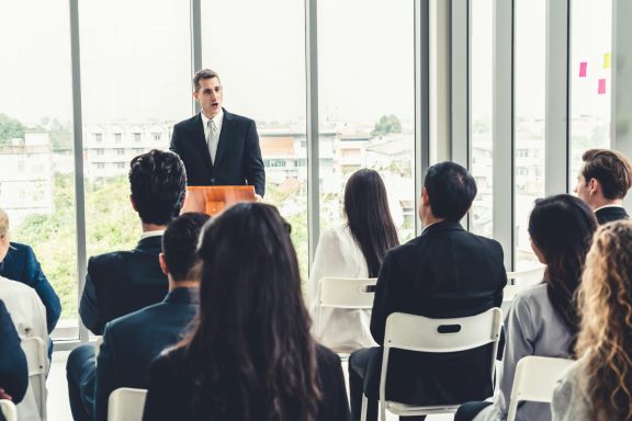 A speaker addresses an audience in a modern conference setting.